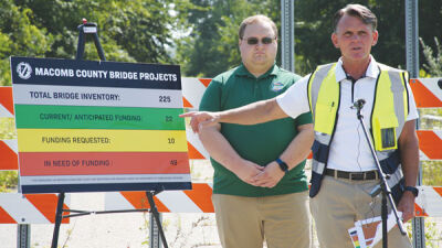  Macomb County Executive Mark Hackel points at a sign outlining the state of bridge construction projects across the county. Lenox Township Supervisor Anthony Reeder Jr. stands behind him. 